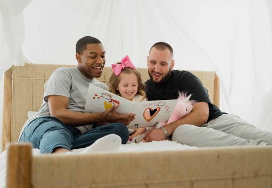 An image of two men smiling and sitting on a bed with their daughter, reading her a story.